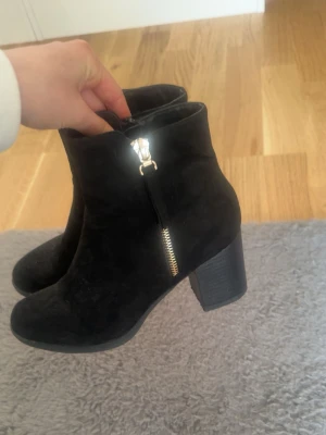 Svarta mockastövletter med klack - Snygga svarta stövletter i mocka med grov klack och dragkedja i silver på sidan. Klassisk rund tå och stilren design som passar till många outfits. Perfekta för dig som vill ha både stil och komfort.