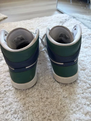 Air Jordan 1 Mid grön/blå/vit - Snygga Air Jordan 1 Mid sneakers i grönt, blått och vitt läder. Klassisk Jordan Wings-logga på sidan och blå Swoosh. Skorna har vita snören och en vit/blå sula. Perfekta för dig som gillar streetwear och vill sticka ut med färg.