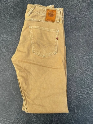 Beige jeans från Replay - Snygga beige jeans från Replay med klassisk femficksdesign och raka ben. Jeansen har läderpatch med Replay-logga bak i midjan och diskret röd detalj på bakfickan. Tillverkade i bomull för en skön känsla och avslappnad look. I storlek W30 L34