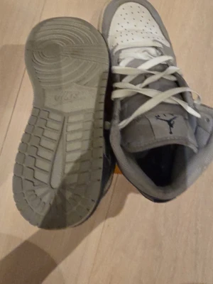 Gråvita Air Jordan 1 Low sneakers - Snygga Air Jordan 1 Low sneakers i grått och vitt med klassisk Jumpman-logga på plösen. Ovandelen är i läder och textil, med perforerad tåbox och vita skosnören. Yttersulan är grå och har bra grepp. Perfekta för dig som gillar streetwear och sportig stil.