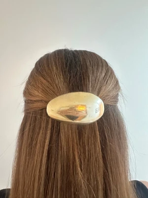 Guldig hårklämma i oval form - Snygg hårklämma i guldig färg med en blank, oval design. Perfekt för att hålla håret på plats och ge en stilren touch till din look. Klämman är tillverkad i plast med metallisk finish och har en enkel men trendig känsla.