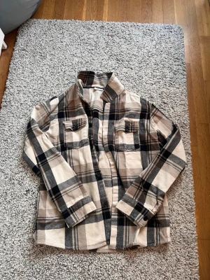 Beige och svart rutig overshirt Kappahl - Overshirt från Kappahl i beige och svart rutigt mönster. Skjortan har två bröstfickor med lock, klassisk krage och knäppning framtill. Tillverkad i mjukt, flanell-liknande material som är perfekt för lager på lager.