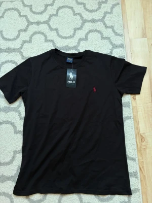 Svart t-shirt från Polo Ralph Lauren - Svart t-shirt från Polo Ralph Lauren med klassisk rund hals och röd broderad logga på bröstet. T-shirten är kortärmad och har en stilren, enkel design som passar till det mesta. Tillverkad i mjuk bomull för en skön känsla.