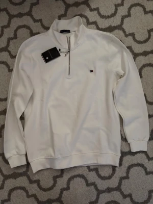 Vit halvzip tröja Tommy Hilfiger - Snygg vit tröja från Tommy Hilfiger med halvzip och ståkrage. Liten broderad logga på bröstet och ribbade muddar vid ärmslut och nederkant. Perfekt till jeans eller chinos för en clean look.