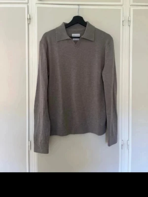 Stickad grå pikétröja These Glory Days - Snygg stickad pikétröja i beige från These Glory Days, tillverkad i mjuk superior merinoull. Tröjan har klassisk krage och v-ringning, perfekt för en stilren och avslappnad look.