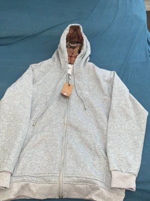Grå zip-hoodie med mönstrat foder - Snygg grå hoodie med dragkedja från Burberry. Utsidan är enkel och stilren, medan insidan av huvan har det klassiska Burberry-rutiga fodret i beige, svart och rött. Perfekt för dig som gillar streetwear med en lyxig touch. Huva med dragsko och mjukt material.