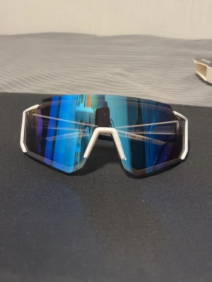 Sportiga vita solglasögon med blå spegelglas - Snygga solglasögon med vit ram och stora blå spegelglas. De har en sportig, futuristisk design med kantiga former och bred båge. Perfekta för dig som vill sticka ut och skydda ögonen mot solen med stil.