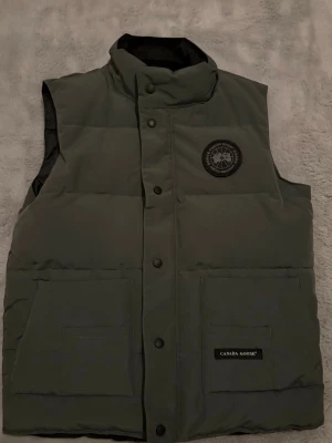Mörkgrön dunväst från Canada Goose - Snygg mörkgrön dunväst från Canada Goose med hög krage, två stora framfickor och svart logotyp på bröstet. Västen har tryckknappar framtill och är fodrad med dun för extra värme. Perfekt för lager-på-lager och streetstyle.