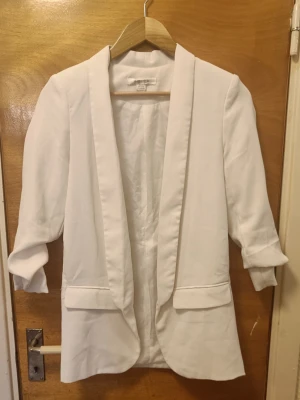 Vit kavaj med upprullade ärmar - Stilren vit kavaj med upprullade ärmar och två fickor framtill. Kavajen har en rak passform och öppen front utan knappar. Perfekt för en trendig och fräsch look. Märket är Amisu och materialet känns mjukt och lätt.