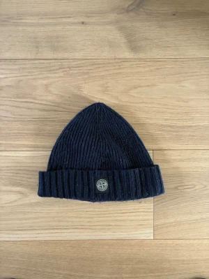 Mörkblå ribbstickad mössa Stone Island - Mörkblå ribbstickad mössa från Stone Island med uppvikt kant och den klassiska logotypen framtill. Tillverkad i ett mjukt och värmande material som passar perfekt för kyliga dagar. Enkel och stilren design som funkar till det mesta.