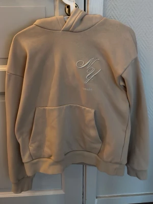 Beige hoodie - Snygg beige hoodie från H&M med broderad logga på bröstet. Hoodien har en stor känguruficka framtill, ribbade muddar och en klassisk huva. Perfekt för en chill och avslappnad stil. Materialet känns mjukt och skönt mot huden.