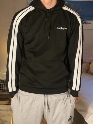 Svart hoodie med vita ränder - Svart hoodie med vita ränder längs ärmarna och texten 'Los Angeles' på bröstet. Klassisk huva med dragsko och ribbade muddar. Sportig och stilren design, perfekt för en avslappnad look.