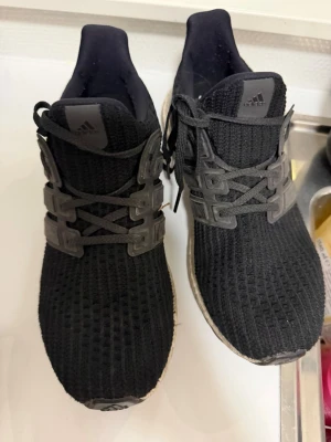 Svarta Adidas Ultraboost sneakers - Svarta Adidas Ultraboost sneakers med stickad ovandel och klassiska tre ränder i plast på sidorna. Skorna har en vit Boost-sula och svarta skosnören. Perfekta för dig som gillar sportig och stilren look.