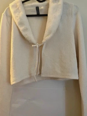 Vit ribbad kofta med fuskpäls - Superfin vit ribbad kofta från H&M med mjuk fuskpäls runt kragen och knytband framtill. Kort modell med långa ärmar, perfekt för lager på lager. Materialet är mjukt och bekvämt, och kragen ger en extra mysig känsla.