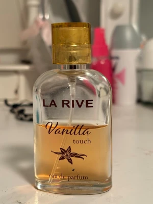 La Rive Vanilla Touch parfym - La Rive Vanilla Touch eau de parfum med en söt och varm vaniljdoft. Flaskan är genomskinlig med guldfärgat lock och har elegant text och en vaniljblomma på framsidan. Perfekt för dig som gillar mjuka och söta dofter. Halv använd som ni ser.
