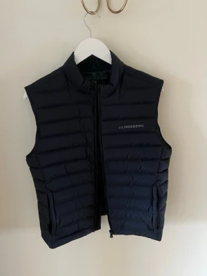 Mörkblå dunväst från J.Lindeberg - Snygg mörkblå dunväst från J.Lindeberg med diskret logga på bröstet. Västen har storlek M men passar nog snarare som S. Väldigt gott skick. 