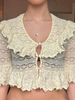 Ljus gul topp med volang - Supersöt croppad blus i gul spets med volangdetaljer och knappar framtill. Blusen har korta ärmar och v-ringning, perfekt för dig som gillar romantisk och vintage-inspirerad stil. Materialet är spets och toppen har en loose passform.