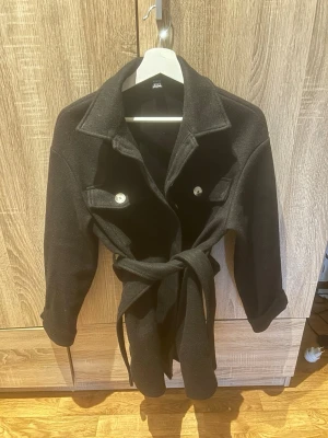 Svart kappa med knytskärp - Stilren svart kappa med bred krage och knytskärp i midjan. Två bröstfickor med knappar framtill ger en cool touch. Perfekt för dig som vill ha en klassisk men trendig look. Materialet ser ut att vara ullblandning.