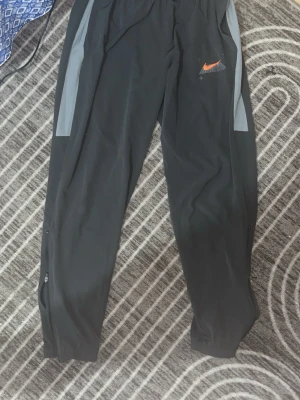 Svarta träningsbyxor från Nike - Säljer ett par svarta träningsbyxor från Nike med gråa detaljer längs sidorna och en orange Nike-logga på låret. Byxorna har dragkedjor vid bensluten och är tillverkade i ett lätt och funktionellt material, perfekta för träning eller löpning.