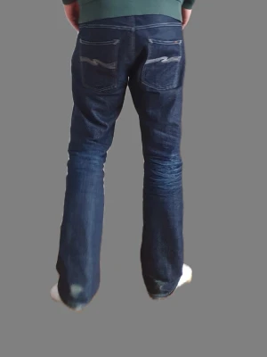 Nudie Jeans - Säljer ett par mörkblå jeans från Nudie Jeans 2001 med klassisk femficksmodell och kontrastsömmar. Jeansen har raka ben, snygga slitningar framtill och broderade bakfickor. Tillverkade i kraftigt denimtyg som ger en cool och tidlös look.