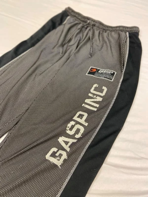 Svarta mjukisbyxor från GASP - Snygga svarta mjukisbyxor från GASP med gråmelerade partier och vita detaljer. Byxorna har dragsko i midjan och logga på benet. Perfekta för träning eller chill. Sköna och stilrena med loose fit.