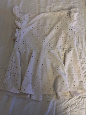 Vit topp  med volang och knyt - Söt vit topp med broderade hålmönster, volangdetaljer och puffärmar. Klänningen har v-ringning med knyt framtill och är tillverkad i ett lätt bomullstyg. Perfekt för sommarens varma dagar.