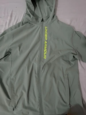 Grå hoodie från Under Armour - Snygg grå hoodie från Under Armour med dragkedja och huva. Framsidan har ett gult Under Armour-tryck längs dragkedjan och diskret logga på bröstet. Tillverkad i ett mjukt och stretchigt material som passar perfekt till träning eller chill.