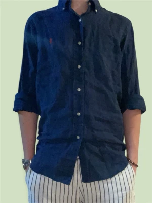 Ralph Lauren skjorta - Klassisk mörkblå skjorta från Ralph Lauren med knappslå och vit knappdetalj. Skjortan har lång ärm och ikonisk röd logga broderad på bröstet. Tillverkad i mjuk bomull och har en normal passform som funkar till många olika stilar.