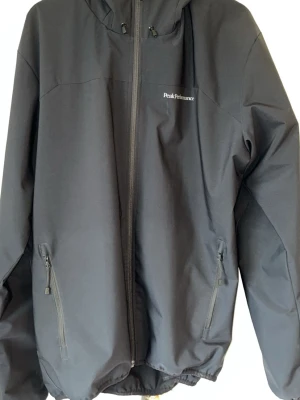 Svart vindjacka från Peak Performance - Snygg svart vindjacka från Peak Performance med huva och dragkedja framtill. Jackan har två sidofickor med dragkedja och diskret logga på bröstet. Tillverkad i ett slitstarkt material som står emot vind. Perfekt för dig som vill ha en stilren och funktionell jacka.