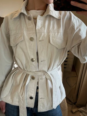 Beige vårjacka - storlek S - Jeansjacka i 100% bomull med knytband i midjan!  - Lindex  Två diskreta fickor på sidorna och två låssas fickor på bröstet, silvriga knappar för stängning  Storlek S Aldrig använd☺️