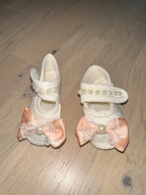 Söta babyskor med rosett och pärlor - Ett par vita babyskor med dekorativa aprikosfärgade rosetter framtill och pärldetaljer på kardborrebandet. Skorna är i tygmaterial och har en mjuk sula, perfekta för små fötter. Passar till dop eller speciella tillfällen.