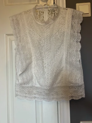 Vit spetsblus från Only - Supersöt vit blus från Only i spets med detaljerade broderier och vågiga kanter. Ärmlös modell med hög hals och rak passform. Perfekt att styla med jeans eller kjol för en trendig look.