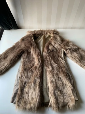 Brun fuskpäls kappa med foder - Snygg brun fuskpäls kappa med mjukt beige foder. Kappan har lång ärm och en rak, något oversized passform. Pälsen är fluffig och ger en lyxig känsla, perfekt för att höja din outfit. Inga synliga knappar eller dragkedja framtill.