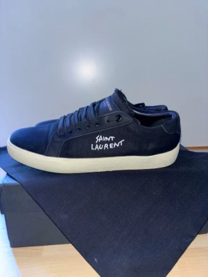 Svarta Saint Laurent sneakers - Snygga svarta sneakers från Saint Laurent med handskriven logga på sidan. Skorna har ovandel i canvas och detaljer i skinn, samt en vit platt gummisula. Klassisk låg modell med snörning och diskreta Saint Laurent-detaljer på plös och sula. Använda fåtal gånger