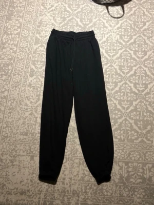 Svarta mjukisbyxor med dragsko - Säljer ett par svarta mjukisbyxor med resår i midjan och dragsko för skön passform. Byxorna har även resår vid bensluten och är gjorda i ett mjukt, bekvämt material. Perfekta för chill dagar hemma eller när du vill vara extra bekväm.