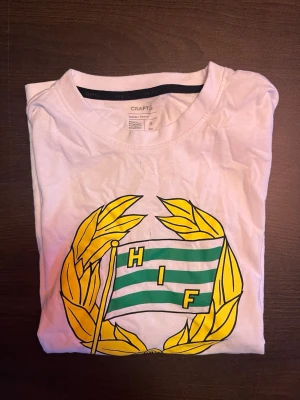 Vit Hammarby T-shirt från Craft - Vit t-shirt från Craft med stort Hammarby IF-tryck på bröstet. T-shirten har klassisk passform, korta ärmar och är gjord i mjuk bomull. Perfekt för dig som vill visa ditt stöd för Hammarby på ett stilrent sätt.
