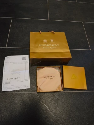 Burberry bälte med kvitto - Snyggt svart bälte från Burberry med klassiskt rutigt mönster på insidan och en stor silverfärgad 'TB'-logga som spänne. Bältet har exklusiv känsla och levereras med originalpåse box och kvitto..