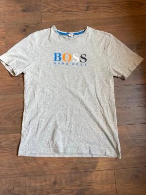 Grå Hugo Boss t-shirt med tryck - Snygg grå t-shirt från Hugo Boss med färgglatt BOSS-tryck i blått, orange, vitt och mörkblått på bröstet. Klassisk passform och rund halsringning. Perfekt för en stilren och avslappnad look. T-shirten har någon fläck som ni ser på bilderna.