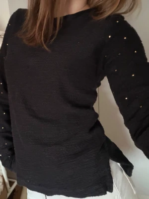 Svart tröja med små nitar - Svart långärmad tröja med struktur och små guldiga nitar på ärmarna. Tröjan har rund halsringning och en något lös passform. Perfekt för dig som vill ha en enkel men ändå lite edgy look.