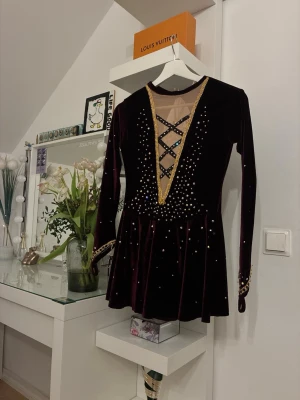 Vinröd sammetsklänning med paljetter - Fin konståkningsklänning i lila sammet med många glittrande stenar. Klänningen har en elegant design med mesh och korsande band fram och bak samt guldfärgade detaljer vid halsen och ärmarna. Kjolen faller fint och rör sig vackert på isen.