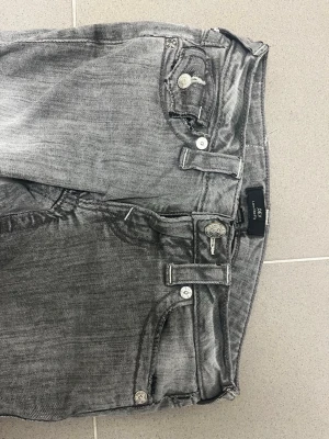 Grå jeans från True Religion - Säljer ett par grå tvättade jeans från True Religion med snygga detaljer som dekorativa sömmar och silverfärgade knappar. Jeansen har klassisk femficksmodell och en unik ficka med lock bak. Perfekta för dig som gillar streetwear och vill sticka ut.