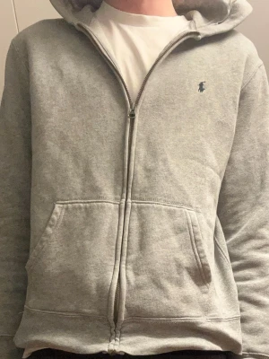 Grå zip hoodie från Polo Ralph Lauren - Snygg grå hoodie med dragkedja från Polo Ralph Lauren. Klassisk design med huva, två stora fickor framtill och den ikoniska lilla loggan broderad på bröstet. Tillverkad i mjukt sweatshirtmaterial som är perfekt för chill dagar.