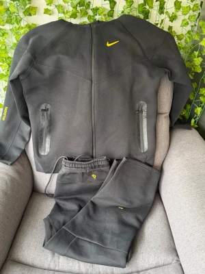 Nike svart tracksuit med gula detaljer - Svart tracksuit från Nike med gula loggor och detaljer. Setet består av en ziptröja med två dragkedjefickor och matchande byxor med resår i midjan. Materialet är mjukt och stretchigt, perfekt för träning eller chill. Snygg design med diskreta sömmar och tryck.