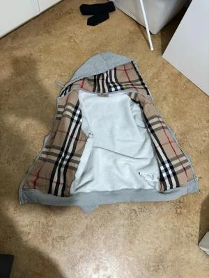Burberry hoodie - Säljer en grå hoodie från Burberry med det klassiska beige, röda och svarta rutiga fodret på insidan. Hoodien har dragkedja och huva med dragsko. Perfekt för dig som vill ha en stilren och lyxig look med ikoniskt mönster.
