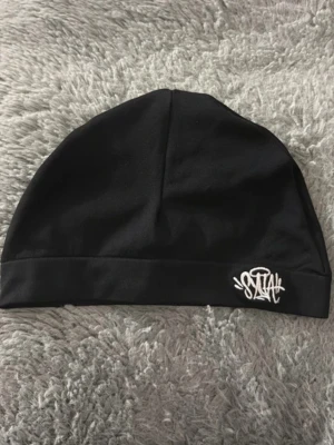 Svart mössa från Sztah - Snygg svart mössa från Sztah med vit broderad logga framtill. Tillverkad i mjukt och stretchigt tyg som sitter skönt på huvudet. Perfekt accessoar för att komplettera din streetwear-look.