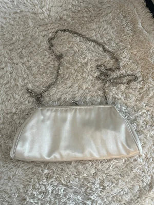 Vit satinkuvertväska med kedja - Stilren vit kuvertväska i satin med silverfärgad kedja som axelrem. Väskan har en klassisk avlång form och stängs med ett litet metallspänne upptill. Några småfläckar, syns på bilderna! 