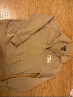 Beige hoodie från Polo Ralph Lauren - Snygg beige hoodie från Polo Ralph Lauren med halv dragkedja och vit logga på bröstet. Tillverkad i mjuk bomullsmix och har en klassisk känguruficka framtill. Perfekt för en avslappnad och stilren look.
