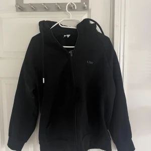 Svart hoodie med dragkedja från ICANIWILL - Svart hoodie från ICANIWILL med dragkedja framtill och justerbar huva. Diskret logga på bröstet och ribbade muddar vid ärmslut och nederkant. Perfekt för chill eller träning. Mjuk bomullsblandning för skön känsla.