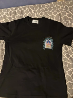 Svart Casablanca t-shirt med tryck - Snygg svart t-shirt från Casablanca med ett färgglatt tryck på bröstet som föreställer en byggnad och texten 'Avenida Casablanca'. Klassisk passform och rund hals. Perfekt för dig som vill ha en stilren men unik look.