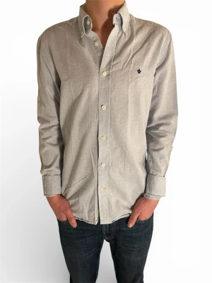 Ljusgrå skjorta från Morris - Klassisk ljusgrå skjorta från Morris med diskret rutmönster och mörkblå broderad logga på bröstet. Skjortan har knappar framtill,  Nypris:1300kr, säljer min för endast 399kr